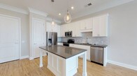 Located at 1414 Broadview Ave in Columbus, Ohio, this single-floor residence offers a thoughtfully designed living environment with contemporary finishes and a clean, modern aesthetic. The kitchen serves as a centerpiece of the home, showcasing crisp white shaker-style cabinetry that extends to the upper walls, complemented by a neutral gray subway tile backsplash that adds texture and depth. A large central island with a smooth dark gray countertop provides generous workspace and casual seating, while pendant lights with teardrop glass shades hang above, casting warm ambient light. Stainless steel appliances — including a French door refrigerator, built-in microwave, electric range, and dishwasher — complete the fully equipped kitchen. Light-toned hardwood flooring runs throughout, contributing to a bright and airy feel. Crown molding along the upper perimeter of the kitchen adds an elegant architectural detail. Recessed lighting further enhances the overall illumination of the space, creating a welcoming and functional atmosphere. The neutral color palette of whites, grays, and warm wood tones gives the home a timeless, move-in-ready appeal suited for a variety of interior styles.