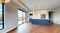 This thoughtfully designed single-floor residence blends contemporary finishes with warm, inviting details throughout. The open-concept kitchen and living area forms the heart of the home, anchored by striking herringbone-patterned hardwood floors that flow seamlessly from room to room. The chef's kitchen is a standout, featuring floor-to-ceiling white shaker cabinetry, a herringbone-tile backsplash, a large slate-blue island topped with quartz countertops, brass hardware and fixtures, pendant lighting with gold accents, and a professional-style range with matching slate-blue finish. Stainless steel appliances complete the culinary space. The living room offers a cozy gas fireplace flanked by built-in shelving, a tray ceiling with recessed lighting, and a modern chandelier. Large black-framed windows and a sliding glass door flood the space with natural light and provide views of the surrounding treeline and outdoor deck. The bedrooms feature plush carpeting and generous black-framed windows with outdoor views, while a walk-in closet with built-in shelving adds organizational convenience. The bathroom is appointed with slate-blue cabinetry, gold hardware, and herringbone luxury vinyl plank flooring, tying the design language of the home together cohesively.