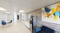 This single-floor common area serves as the central hub of a residential building, combining functionality with a clean, contemporary aesthetic. The lobby is anchored by a textured stone-clad accent wall that adds warmth and visual depth to the space. A comfortable navy blue sofa and a bold, colorful abstract canvas create an inviting lounge area, complemented by a decorative geometric mirror and a sleek console table. A water dispenser and thoughtful furnishing details complete the welcoming reception zone.

An extensive bank of individual mailboxes lines the hallway wall, providing convenient mail access for residents. The corridor extends with light-toned tile flooring underfoot and a bright drop ceiling with recessed panel lighting, ensuring the passageway feels open and well-illuminated throughout.

A glazed fitness center is accessible from the main lobby area, enclosed behind full-height glass walls that offer clear views of the exercise equipment inside, including cardio machines. Natural light filters in through glass exterior doors that open to a green outdoor area, enhancing the sense of openness. A small round table with blue chairs offers a casual seating spot within the common area. The overall design is cohesive, emphasizing light tones, modern furnishings, and practical amenities for residents.