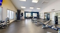 Located at 6200 Rime Village Dr NW in Huntsville, Alabama, this fitness center offers a single-floor exercise room designed to support a comprehensive workout experience. The space features soaring vaulted ceilings that lend an open, airy feel, complemented by large recessed panel lighting and multiple windows fitted with blinds that allow ample natural light while maintaining privacy. The flooring throughout is durable dark rubber, ideal for high-traffic fitness use.

The room is well-equipped with a wide variety of strength and conditioning equipment, including cable machines, leg extension and curl stations, a vertical climbing machine, multiple adjustable weight benches, a barbell rack, and a full dumbbell rack. A wall-mounted mirror along one side enhances the sense of space and allows for form checks during workouts. Two wall-mounted flat-screen TVs provide entertainment and keep users engaged during their sessions.

A bold navy blue accent wall adds a modern, energetic visual element to the otherwise neutral white interior. A set of blue lockers near the glass-paned entry door offers convenient storage for personal belongings. The glass door connects the fitness center to an adjacent area, providing easy access. Overall, the space is thoughtfully laid out to maximize functionality while maintaining a clean, professional atmosphere conducive to focused training.