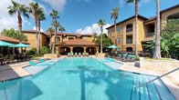 This outdoor amenity space showcases a resort-style swimming pool as its centerpiece, with crystal-clear turquoise water that reflects the open sky above. The pool area is generously proportioned, offering ample room for both leisurely swimming and relaxation. Surrounding the pool, a well-maintained deck is furnished with lounge chairs and seating, providing comfortable spaces to unwind in the sun.

A covered cabana or pavilion serves as a focal point adjacent to the pool, featuring an open-air design with arched colonnades and a terra cotta tile roof, consistent with the Mediterranean or Spanish Colonial architectural style carried throughout the complex. Teal patio umbrellas add a vibrant pop of color and functional shade throughout the deck area.

Tall, mature palm trees are strategically placed around the pool, enhancing the tropical, resort-like ambiance and providing natural shade. The surrounding multi-story building features warm golden-yellow stucco exteriors, red tile roofing, and private balconies, further reinforcing the cohesive Mediterranean aesthetic. The overall atmosphere is bright, inviting, and evocative of a luxury resort setting.