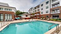 Located at 1813 Sir Tyler Dr in Wilmington, North Carolina, this community amenity and leasing center is defined by a striking combination of warm brick masonry and clean white lap siding, lending the building a modern coastal aesthetic. The exterior entry features a covered portico with arched brick detailing, wall-mounted lantern-style sconces, and large glass-panel doors that provide an inviting, transparent view into the interior. The entryway is finished with a smooth concrete floor and a branded welcome mat, establishing a polished first impression.

Inside, exposed brick accent walls add character and a sense of permanence to the interior spaces, complementing the overall design language carried throughout the building. The layout connects seamlessly to the outdoor amenity areas through gated access points, maintaining both security and visual continuity.

The standout outdoor feature is a generously sized resort-style swimming pool, surrounded by a spacious stone-paved deck furnished with lounge chairs, vibrant orange and teal outdoor seating, and shade umbrellas. A covered pavilion with a mounted outdoor television provides an additional gathering space. Palm trees and lush landscaping frame the courtyard, while the multi-story residential buildings surrounding the pool create a cohesive, enclosed community atmosphere. The overall ambiance is relaxed, upscale, and well-suited to the warm Wilmington climate.