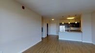 This single-floor space showcases a thoughtfully designed open-concept layout that seamlessly connects the living area to the kitchen. Warm hardwood-style flooring extends throughout, adding a cohesive and welcoming feel to the interior. The neutral-toned walls are complemented by clean white baseboards, creating a bright and airy atmosphere.

The kitchen is a standout feature, equipped with dark cabinetry, stainless steel appliances, and a built-in microwave. A light-toned stone countertop breakfast bar serves as both a functional workspace and a casual dining area. Pendant lighting fixtures hang above the kitchen island, providing focused illumination and a contemporary design touch.

The open living area offers generous space, with recessed and flush-mount ceiling lighting ensuring the area is well-lit. Two interior doors visible from the main space lead to additional rooms or storage areas, suggesting a practical and functional floor plan. The overall ambiance is modern and clean, with a neutral palette that provides a versatile backdrop well-suited to a variety of interior styles.