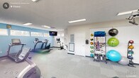 Located at Brookstone Lane in Fayetteville, NC, this dedicated fitness room offers a comprehensive exercise environment thoughtfully designed to support a wide range of workout routines. The single-floor space is organized to maximize functionality, with clearly defined zones for cardio, strength training, and accessory work.

The cardio section features multiple treadmills and stationary bikes, including both upright and recumbent models, positioned along a light blue accent wall with horizontal windows that invite natural light into the room. A wall-mounted flat-screen television provides entertainment during workouts.

A full-length wall of mirrors along one side visually expands the space and aids in form monitoring, complementing the strength training area which includes a cable machine, a weight bench, and additional resistance equipment. Recessed and panel fluorescent lighting ensures consistent, bright illumination throughout.

Along the opposite wall, a well-organized equipment rack holds an array of accessories including medicine balls, resistance bands, exercise balls, and kettlebells, keeping the floor clear and workout-ready. Neutral tile-pattern carpet flooring covers the entire room, offering comfort and durability underfoot.

A fire extinguisher, informational signage, and a workout board round out the practical amenities, making this a fully functional and inviting fitness facility.