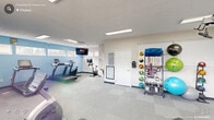 Located at Brookstone Lane in Fayetteville, NC, this dedicated fitness room offers a comprehensive exercise environment thoughtfully designed to support a wide range of workout routines. The single-floor space is organized to maximize functionality, with clearly defined zones for cardio, strength training, and accessory work.

The cardio section features multiple treadmills and stationary bikes, including both upright and recumbent models, positioned along a light blue accent wall with horizontal windows that invite natural light into the room. A wall-mounted flat-screen television provides entertainment during workouts.

A full-length wall of mirrors along one side visually expands the space and aids in form monitoring, complementing the strength training area which includes a cable machine, a weight bench, and additional resistance equipment. Recessed and panel fluorescent lighting ensures consistent, bright illumination throughout.

Along the opposite wall, a well-organized equipment rack holds an array of accessories including medicine balls, resistance bands, exercise balls, and kettlebells, keeping the floor clear and workout-ready. Neutral tile-pattern carpet flooring covers the entire room, offering comfort and durability underfoot.

A fire extinguisher, informational signage, and a workout board round out the practical amenities, making this a fully functional and inviting fitness facility.