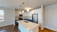 This well-designed single-floor unit offers a clean, contemporary aesthetic with thoughtful finishes throughout. The heart of the home is an open-concept kitchen that seamlessly connects to the living area, creating a spacious and sociable layout. The kitchen is a standout feature, showcasing a striking two-tone cabinetry design with crisp white upper shaker-style cabinets and muted blue-grey lower cabinets, all complemented by bright white quartz countertops. A generously sized central island with a double undermount sink serves as both a functional workspace and a natural gathering point. Stainless steel appliances, including a French door refrigerator, electric range, and over-the-range microwave, complete the kitchen. Warm hardwood-style flooring flows throughout the main living areas, adding a sense of continuity and warmth. Pendant lighting with gold-toned fixtures adds a modern, refined touch above the island and in the living space. The bedroom features plush neutral carpeting and a bright window with blinds that allows for natural light while maintaining privacy. The bathroom is finished with a grey vanity, white countertop, a tub-shower combination, and matching hardwood-style flooring, offering a clean and functional design. Overall, the space presents a fresh, move-in-ready feel with modern finishes and a cohesive color palette.