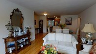 Located at 171 Main St in Madison, NJ, this single-floor residence offers a welcoming and well-connected living environment with a warm, classic aesthetic. The open-concept layout seamlessly connects the living room, dining area, and kitchen, creating a cohesive and functional flow throughout the home. The living room is anchored by a comfortable white sofa, complemented by rich hardwood flooring that extends throughout the main living spaces, adding warmth and continuity. An ornate wooden console with a decorative mirror enhances the traditional character of the space. The dining area, positioned between the living room and kitchen, features a classic round table with seating, framed by natural light filtering in through the windows. The kitchen is equipped with stainless steel appliances, including a full-size refrigerator, set against warm wood cabinetry and accented by a decorative valance above the window. Recessed lighting in the kitchen provides functional illumination, while a breakfast bar area adds versatility for casual dining. A stately grandfather clock and carefully chosen artwork and floral arrangements throughout contribute to the home's inviting, traditional ambiance, making it a comfortable and characterful single-level residence.