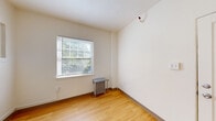 This single-floor space presents a clean and minimalist aesthetic, defined by crisp white walls, warm honey-toned hardwood flooring, and an airy, understated ambiance throughout. The layout encompasses several distinct rooms, each sharing a cohesive design language that emphasizes simplicity and functionality.

The main room is generously proportioned, featuring a large window fitted with horizontal blinds that frames lush greenery outside, allowing natural light to pour in and cast bright, shifting patterns across the hardwood floor. A classic cast-iron radiator sits beneath the window, nodding to the building's character and providing efficient heating. A transom window is also visible, adding an architectural detail that lends a sense of history to the space.

An adjoining room offers the same warm flooring and white walls, with a wall-mounted sink accompanied by a vanity mirror and a lighted fixture above — a practical and neatly integrated feature. Additional rooms maintain the same palette and materials, with white panel doors, clean baseboards, and ample wall space offering flexibility for a variety of uses.

Sunlight streams generously through the windows, illuminating the interiors and enhancing the sense of openness. The overall atmosphere is calm and versatile, making this space well-suited for a range of occupancies or configurations.
