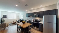 This single-floor unit presents a thoughtfully designed layout with a contemporary aesthetic throughout. The open-concept kitchen and living area form the heart of the space, connected seamlessly by light-toned hardwood flooring that extends across the main living zones. The kitchen is well-appointed with dark espresso cabinetry reaching toward the ceiling, providing ample storage. Stainless steel appliances — including a refrigerator, electric range, over-the-range microwave, and dishwasher — complement the clean white countertops. A butcher block kitchen island with bar seating offers additional prep space and casual dining. Recessed lighting illuminates the kitchen efficiently, while natural light filters in through slatted window blinds. The living area is open and comfortable, featuring neutral walls, a large window that brings in natural light, and hardwood flooring that ties the space together. A dedicated media corner accommodates a television and accent seating. The bedroom is carpeted for comfort, featuring neutral walls, a window with blinds, and sufficient space for a full bedroom suite. The overall ambiance is clean, modern, and functional, with a consistent design language of dark accents against light surfaces.