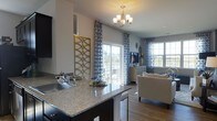This single-floor residence showcases a thoughtfully designed open-concept layout that connects the kitchen and living areas into one cohesive, flowing space. The kitchen features dark cabinetry contrasted by a granite countertop with speckled detailing, a stainless steel sink, and ample counter space — combining both style and functionality. A decorative gold geometric panel adds an artistic accent, complementing the warm, elegant tone of the space.

A chandelier with frosted glass shades serves as a central focal point overhead, casting warm light throughout. Hardwood-style flooring runs continuously from the kitchen into the living area, reinforcing the open and unified feel. The living space is generously sized and bathed in natural light, courtesy of multiple windows and a sliding glass door that leads to an outdoor area, framed by patterned blue and white curtains that add a soft, decorative touch.

The living area accommodates comfortable seating and a media console, creating a welcoming and well-appointed environment. The overall ambiance is bright, modern, and elegantly styled with a cohesive neutral and gold color palette.