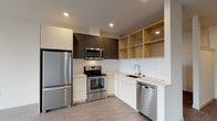 This contemporary single-floor unit showcases a clean, modern design with an open-concept layout and neutral color palette throughout. The kitchen is a standout feature, equipped with a full suite of stainless steel appliances including a refrigerator, electric range with over-the-range microwave, and dishwasher. Cabinetry combines light lower cabinets with dark upper cabinets and open natural wood shelving, all complemented by a white subway tile backsplash and light countertops. The kitchen flows seamlessly into a generously proportioned living area finished with durable wood-grain luxury vinyl plank flooring that extends throughout the unit. Large, floor-to-ceiling windows in both the living and bedroom areas flood the interior with natural light and frame expansive views of the surrounding urban cityscape. The bedroom is well-proportioned, offering a corner window for added light and scenic views. The bathroom features an elongated double-sink vanity with dark wood cabinetry, light quartz countertops, a large wall mirror, and clean white tile flooring, creating a sleek and functional space. Recessed lighting throughout enhances the bright, airy ambiance of the entire unit.