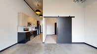 This single-floor unit showcases a compelling blend of industrial and contemporary design, defined by soaring ceilings with exposed concrete and ductwork that lend a loft-like character throughout. Polished concrete floors extend across the open main living area, reflecting light and reinforcing the space's modern aesthetic.

The kitchen is thoughtfully appointed with two-tone cabinetry — light wood upper cabinets paired with dark lower units — set against a classic white subway tile backsplash. Stainless steel appliances, including a refrigerator, range, microwave, and dishwasher, complete the functional layout. Striking black-and-gold pendant lights hang overhead, adding a warm, stylish accent.

A bold black sliding barn door connects the main living area to the bedroom, which features carpeted flooring for comfort, tall ceilings with exposed HVAC elements, and direct access to a private balcony through full-height black-framed glass doors. The balcony brings in generous natural light and outdoor views.

The bathroom is finished in a clean, minimalist style with a dark-framed vanity, white countertop, matte black fixtures, and a full tub-shower combination surrounded by white tile. A large mirror and simple, modern hardware complete the look. The entry corridor features a secure keypad door, providing a polished and practical welcome into the space.