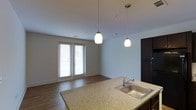 This single-floor space showcases a thoughtfully designed open-concept layout that seamlessly connects the kitchen and living areas. The kitchen features a granite-look countertop island with a stainless steel sink, providing both functionality and a central gathering point. Dark cabinetry adds a rich contrast to the neutral, light-toned walls, while two stylish pendant lights hang overhead, offering focused task lighting and a modern aesthetic touch. The living area is anchored by warm hardwood-style flooring that extends throughout the space, adding a sense of cohesion and warmth. A set of French doors with white blinds serves as the focal point of the living area, allowing an abundance of natural light to fill the room and providing access to an outdoor space. The overall color palette — soft whites, warm wood tones, and dark accents — creates a balanced and welcoming ambiance. The open layout and clean architectural lines make the space feel airy and well-proportioned, ideal for both everyday living and entertaining.