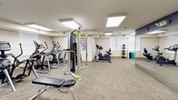 Located at 420 Riverglade Dr in Amherst, Massachusetts, this dedicated exercise room offers a well-appointed fitness facility across a single floor. The space is thoughtfully designed to accommodate a wide variety of workout routines, housing an impressive array of Matrix-brand cardio machines, including treadmills, ellipticals, and stationary bikes, alongside a full complement of strength training equipment such as cable machines, weight benches, and a medicine ball rack. The room is finished with durable, neutral-toned carpet tiles that provide both comfort underfoot and a clean, professional aesthetic. Recessed panel lighting across the ceiling ensures bright, even illumination throughout the workout area. Large wall-mounted mirrors along one side of the room enhance the sense of openness while allowing users to monitor their form during exercise. Windows along the perimeter allow natural light to supplement the interior lighting, contributing to an energizing atmosphere. The space also includes a climate control unit for year-round comfort, a fire safety system, and direct access to an adjacent corridor. Informational postings near the entrance provide guidelines for equipment use. Overall, the facility is practical, efficiently organized, and well-maintained, making it a functional fitness resource.