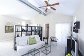 This single-floor residence offers an open-concept studio layout that makes efficient and stylish use of its footprint. The main living area immediately impresses with soaring vaulted ceilings accented by exposed industrial ductwork, giving the space a loft-like character. A ceiling fan provides comfort throughout, while large windows invite generous natural light. A freestanding bookcase unit serves as a functional room divider, subtly delineating the sleeping area from the living space without sacrificing the open feel. Plush carpeting runs through the living and sleeping zones, adding warmth underfoot.

The kitchen is outfitted with warm-toned wooden cabinetry, dark countertops, and black appliances including an electric range and an overhead microwave. Light hardwood flooring in the kitchen and adjoining hallway contrasts nicely with the carpeted living areas, creating a natural transition between spaces.

The bathroom is thoughtfully designed with rich wood-toned vanity cabinetry, a wide gray countertop, an extended mirror, and built-in wall shelving for additional storage. A shower-tub combination with a decorative curtain completes the space. Together, these elements create a cohesive, comfortable residence that balances industrial-modern design with warm, inviting finishes.