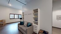 This thoughtfully designed single-floor studio offers a smart, open-concept layout that maximizes every square foot. The main living area serves as the centerpiece of the unit, featuring a built-in Murphy bed system flanked by open shelving and a chest of drawers — a seamless solution that transitions the space between living and sleeping functions. A gray sofa, area rug, and accent chair complete the living zone, which benefits from high ceilings with exposed track lighting that add an industrial-modern character. The kitchen is well-appointed with white shaker-style cabinetry, dark tile backsplash, stainless steel appliances including a gas range, dishwasher, and refrigerator, plus a window that brings in natural light. A separate dining nook provides a defined eating area with a small round table and chairs, complemented by wall-mounted artwork and a wall-unit HVAC system. The bathroom is clean and functional, featuring a white vanity with sink, tile flooring, and a large mirror. Neutral gray walls and dark baseboards run throughout, lending the space a cohesive, contemporary aesthetic ideal for modern living.