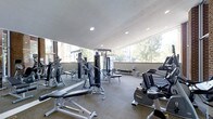 This fitness center offers a comprehensive workout environment designed to accommodate a wide range of exercise needs. The space is flooded with natural light, courtesy of large floor-to-ceiling windows that line the perimeter, offering views of the surrounding greenery and creating an open, airy atmosphere. The smooth, light-toned ceiling is fitted with recessed lighting, ensuring consistent illumination throughout the facility. The flooring transitions between carpet and rubberized mat surfaces, providing appropriate support for both cardio and strength training areas. The strength training section is well-appointed with a variety of cable machines, multi-function weight stations, and adjustable benches — including Matrix-branded equipment — catering to both beginners and experienced athletes. The cardio section features upright bikes, elliptical trainers, and additional aerobic equipment positioned to take advantage of the natural light and window views. Wall-mounted mirrors extend along one side of the space, enhancing the sense of depth and allowing users to monitor their form during workouts. The overall ambiance is clean, modern, and functional, making this fitness center a well-rounded facility for daily training.