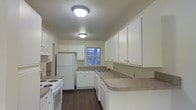 Located at The Brickyard, this single-story space offers a well-organized and functional layout. The kitchen serves as a central feature, designed with ample white cabinetry that provides generous storage along both upper and lower sections. The countertops offer a practical work surface, wrapping around the perimeter to maximize workspace efficiency. White appliances, including a refrigerator and electric range, are seamlessly integrated into the cabinetry layout, contributing to a clean, cohesive aesthetic. A sink is positioned beneath a window, allowing for natural light to brighten the workspace during the day. The dark hardwood-style flooring throughout provides a warm contrast to the lighter cabinetry and walls, adding visual depth to the space. Flush-mount ceiling light fixtures provide consistent overhead illumination throughout. The textured ceiling and neutral-toned walls create a straightforward, practical atmosphere. The kitchen's U-shaped configuration promotes an efficient workflow, making it well-suited for everyday cooking and meal preparation. Overall, the space presents a tidy, functional environment with a neutral palette that lends itself well to a variety of interior styles.