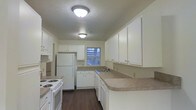 Located at The Brickyard, this single-story space offers a well-organized and functional layout. The kitchen serves as a central feature, designed with ample white cabinetry that provides generous storage along both upper and lower sections. The countertops offer a practical work surface, wrapping around the perimeter to maximize workspace efficiency. White appliances, including a refrigerator and electric range, are seamlessly integrated into the cabinetry layout, contributing to a clean, cohesive aesthetic. A sink is positioned beneath a window, allowing for natural light to brighten the workspace during the day. The dark hardwood-style flooring throughout provides a warm contrast to the lighter cabinetry and walls, adding visual depth to the space. Flush-mount ceiling light fixtures provide consistent overhead illumination throughout. The textured ceiling and neutral-toned walls create a straightforward, practical atmosphere. The kitchen's U-shaped configuration promotes an efficient workflow, making it well-suited for everyday cooking and meal preparation. Overall, the space presents a tidy, functional environment with a neutral palette that lends itself well to a variety of interior styles.