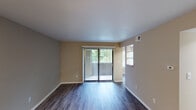 This single-story residence offers a well-organized layout with distinct living, sleeping, kitchen, and bathroom areas. The living room is an open, neutrally toned space finished with dark wood-style vinyl flooring that runs throughout the main areas. A full-width sliding glass door fitted with vertical blinds serves as the room's focal point, drawing in natural light and providing direct access to an outdoor patio with views of surrounding greenery.

Accessible from the living area through an open archway, the galley-style kitchen features white cabinetry with open shelving, light granite-look countertops, a stainless steel sink, and black appliances including a refrigerator, range, and dishwasher. A louvered closet door at the end of the kitchen provides additional storage.

The bedroom is a generously sized room finished with plush carpet and neutral beige walls. A window with horizontal blinds allows natural light and outdoor views. Two louvered closet doors provide ample storage, and ceiling vents are positioned for efficient climate control throughout.

The bathroom features a clean white vanity with under-cabinet storage, a framed mirror, and a well-lit overhead fixture, providing a functional and tidy space. Overall, the unit presents a clean, move-in-ready aesthetic with a consistent neutral palette throughout.