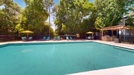 Located at 330 W Fort Morgan Rd in Gulf Shores, Alabama, this outdoor pool area offers a well-appointed recreational retreat designed for relaxation and leisure. The centerpiece is a large, rectangular swimming pool with clear turquoise water, complemented by a wide surrounding concrete deck that provides ample space for sunbathing and socializing. Numerous teal lounge chairs are arranged along the pool's perimeter, offering comfortable spots for guests to unwind under the sunny Alabama sky. Yellow umbrellas are positioned throughout the deck, providing shaded seating areas for those seeking relief from the sun. A wood-frame pergola anchors one corner of the pool area, adding a charming architectural accent and additional shaded gathering space. The pool is enclosed by a black iron fence, ensuring safety and privacy. A single-story pool house with a covered porch, multiple windows, and French-style doors sits adjacent to the pool, offering convenient access to amenities. The entire space is beautifully framed by mature trees and lush greenery, creating a serene, park-like atmosphere. Depth markers are visible along the pool's edge, and clear signage is posted throughout the area for guest safety and guidance.