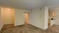 This single-floor space presents a straightforward, functional layout centered around a main living area finished with wall-to-wall beige carpeting that adds a sense of warmth underfoot. The room is defined by smooth, neutral-toned walls painted in a soft off-white, creating a clean and versatile backdrop suitable for a variety of interior styles. The standard ceiling height provides a comfortable, intimate atmosphere throughout the space.

Built-in HVAC floor vents and wall-mounted thermostat and ventilation controls are visibly integrated into the walls, indicating a centralized climate control system. Multiple electrical outlets are positioned at regular intervals along the baseboards, offering practical convenience throughout the room.

A doorway at the far end of the main area leads into a connecting hallway or secondary room, with a hint of additional doors visible beyond, suggesting further rooms or storage access. A narrow vertical window near this entryway introduces a modest amount of natural light into the transitional area. On the left side of the room, a set of what appears to be closet or storage doors adds additional functionality to the space. Overall, the layout is simple, open, and move-in ready.