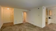 This single-floor space presents a straightforward, functional layout centered around a main living area finished with wall-to-wall beige carpeting that adds a sense of warmth underfoot. The room is defined by smooth, neutral-toned walls painted in a soft off-white, creating a clean and versatile backdrop suitable for a variety of interior styles. The standard ceiling height provides a comfortable, intimate atmosphere throughout the space.

Built-in HVAC floor vents and wall-mounted thermostat and ventilation controls are visibly integrated into the walls, indicating a centralized climate control system. Multiple electrical outlets are positioned at regular intervals along the baseboards, offering practical convenience throughout the room.

A doorway at the far end of the main area leads into a connecting hallway or secondary room, with a hint of additional doors visible beyond, suggesting further rooms or storage access. A narrow vertical window near this entryway introduces a modest amount of natural light into the transitional area. On the left side of the room, a set of what appears to be closet or storage doors adds additional functionality to the space. Overall, the layout is simple, open, and move-in ready.