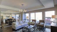 This single-floor space presents an open and thoughtfully designed living area that seamlessly connects to an adjacent dining space. The living room is anchored by a contemporary chandelier with frosted cylindrical shades, suspended above a neutral-toned seating arrangement consisting of a plush sofa and a pair of coordinating wingback chairs. A patterned area rug in cool blues and grays defines the seating zone and adds visual interest underfoot. A rectangular coffee table with a sleek metal frame serves as the functional centerpiece of the arrangement.

Large windows along the exterior wall flood the space with natural light, creating a bright and airy atmosphere throughout. The walls are finished in a warm greige tone that complements the neutral furnishings, while crisp white tray ceilings add architectural dimension and a sense of vertical space. A floor lamp with a classic drum shade provides additional ambient lighting. In the background, the dining area is visible, featuring a dark-toned table and chairs alongside additional natural light from a glass door, reinforcing the open, connected flow of the overall layout.