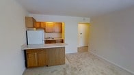 This single-floor space offers a functional and comfortable layout designed for everyday living. The kitchen is a standout feature, equipped with warm honey-toned wood cabinetry that provides ample storage space both above and below the countertops. A white refrigerator and a double sink contribute to the kitchen's practicality, while a peninsula with a light-colored countertop extends outward to create a natural breakfast bar, ideal for casual dining or additional prep space. The kitchen opens seamlessly into the living area, creating an open-concept feel that enhances the sense of spaciousness. Soft, neutral-toned carpeting covers the living area floor, adding warmth and comfort underfoot. The walls are finished in a clean, light cream tone that helps reflect light and contributes to a bright, airy ambiance throughout the space. A doorway visible from the main living area leads to an entryway or additional room, suggesting a thoughtful flow between spaces. The transition between the tiled entryway and the carpeted living area is clearly defined, adding a subtle design distinction. Overall, the space presents a clean, well-maintained interior with a practical layout suited for comfortable day-to-day living.