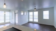 This single-floor residence offers a clean, contemporary design centered around an open-concept living and kitchen layout. Dark hardwood-style flooring runs throughout the main living areas, providing a warm contrast against the crisp white walls and light-toned ceilings. The living area is accented by a ceiling fan with integrated lighting, adding both function and a casual charm to the space.

The kitchen is well-appointed with dark espresso cabinetry, stainless steel appliances including a full-size refrigerator, a black electric range, and granite countertops that extend into a peninsula-style breakfast bar. Pendant lighting hangs above the kitchen area, complemented by track lighting that highlights the cabinetry. Multiple large windows with horizontal blinds flood the kitchen and adjacent dining nook with generous natural light, creating an airy and inviting atmosphere.

The living area opens toward multiple windows and a sliding glass door, offering outdoor views and further enhancing the brightness of the space. A second ceiling fan is positioned in what appears to be a secondary living or dining area, maintaining comfortable airflow. Doorways lead to additional rooms, suggesting a thoughtful and functional floor plan. Overall, the space presents a modern, move-in-ready feel with stylish finishes throughout.