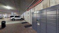 This covered parking garage provides a functional and well-equipped space designed to serve the practical needs of building occupants. The structure features exposed concrete ceilings with prominent red fire suppression piping running overhead, complemented by concrete masonry block walls that give the space a sturdy, utilitarian character. The flooring is finished in classic brick pavers, adding a touch of texture and durability underfoot.

A standout feature of this garage is the Amazon Hub package locker system, a bank of secure, self-service lockers mounted along the wall that allows residents or tenants to retrieve deliveries at their convenience. Adjacent to the lockers is a clearly marked elevator entrance, providing direct and easy access to the building's upper levels.

The garage offers designated parking bays along the interior, accommodating standard-sized vehicles with ease. Overhead lighting illuminates the main travel lanes and key access points, ensuring safe navigation throughout the space. The combination of secure package retrieval, elevator access, and covered parking makes this garage a highly practical and convenient facility for everyday use.