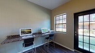 This single-floor business center is a dedicated workspace designed to meet the productivity needs of its users. The room features a continuous wall-mounted countertop with a dark granite-style surface, providing ample desk space for multiple workstations equipped with desktop computers and a printer. Sleek, modern task chairs offer comfortable seating at each station.

The space is finished with warm, two-tone painted walls in muted green and tan hues, complemented by rich, dark hardwood-style flooring that lends a polished, professional feel. Natural light fills the room through large, grid-paned windows that offer views of a landscaped outdoor courtyard and surrounding buildings, creating a bright and welcoming atmosphere. A glass-paneled entry door further enhances the flow of natural light into the space.

A cork bulletin board mounted on the wall serves as an informational hub for notices and announcements. The room also includes network infrastructure, with a wall-mounted router ensuring reliable connectivity for all workstations. Clean white baseboards and a straightforward, uncluttered layout contribute to the room's functional and organized character. Overall, the space provides a quiet, well-equipped environment well-suited for focused work tasks.