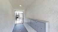 This single-story common area serves as the primary entryway for the building, combining security and practicality in a compact, efficiently designed corridor. The space is defined by its textured stucco walls finished in a neutral white tone, complemented by a matching popcorn-textured ceiling that contributes to the cohesive aesthetic throughout. A recessed ceiling light provides consistent overhead illumination, ensuring the corridor remains well-lit at all times.

A prominent feature of this entryway is the extended bank of individual metal mailboxes lining one wall. The mailboxes are finished in a muted blue-grey tone and include a variety of unit compartments, offering residents a centralized and organized mail retrieval area. The corridor leads to a wrought-iron security gate at the far end, which allows natural light to filter into the space while maintaining controlled access to the building's exterior. Beyond the gate, an outdoor area is partially visible, suggesting a courtyard or pathway. The floor is covered in a durable grey carpet, adding a modest degree of warmth to the otherwise utilitarian corridor.