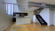 This two-floor loft-style space blends industrial character with modern finishes to create a dynamic and functional living environment. The main level showcases an open-concept layout where the kitchen flows seamlessly into the living area, fostering a sense of spaciousness and connectivity. The kitchen is appointed with dark espresso cabinetry, stainless steel appliances including a refrigerator and microwave, and a central island with a light granite countertop that provides additional prep and seating space. A pendant chandelier with multiple gooseneck lights hangs above the island, adding a stylish focal point. Overhead, an exposed cylindrical metal duct runs prominently across the ceiling, lending the space an authentic industrial aesthetic that complements the overall loft character. The main level is grounded by warm, light-toned hardwood flooring that extends throughout, adding natural warmth to the open environment. A bold black metal staircase with clean lines rises to the upper floor, serving as both a functional and architectural statement piece. The upper level overlooks the main floor through a metal railing, enhancing the vertical sense of space. The high ceilings and open sightlines across both levels contribute to an airy, loft-inspired atmosphere throughout.