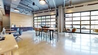 Located at 525 Lincoln St in Boston's Allston neighborhood, this single-floor commercial space offers a thoughtfully designed open-plan layout spanning a generous footprint. The interior showcases an industrial-chic aesthetic, characterized by exposed ductwork and mechanical systems overhead, polished concrete floors, and structural concrete columns that lend the space an authentic, loft-like character.

Natural light floods the interior through dramatic floor-to-ceiling glass garage doors along the street-facing wall, providing sweeping views of the surrounding streetscape and creating a bright, energizing atmosphere throughout. The space is organized into distinct yet fluid zones: a casual lounge area features a sculptural swivel chair on a patterned area rug, set against a warm wood-paneled accent wall adorned with circular textured art pieces. A communal dining zone offers both a live-edge high-top table with bar seating and a round table surrounded by wire-frame chairs.

A built-in cabinetry and kitchenette area provides practical amenities along one wall, while a semi-enclosed room visible through glass partitions offers a more intimate setting with pendant lighting. Curated artwork, pendant and track lighting, and a wall-mounted display screen round out the refined yet functional design, making this a highly versatile space well-suited for collaboration, gathering, or hospitality.