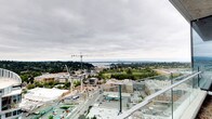 This rooftop terrace is positioned at a commanding elevation, providing an expansive, unobstructed panorama that stretches across a dynamic urban landscape. Glass panel railings line the perimeter, ensuring safety while preserving uninterrupted sightlines in every direction. The open-air design allows occupants to take in the full breadth of the surroundings, from active street-level development and mid-rise buildings in the foreground to a dense tree canopy and a shimmering body of water in the distance. On clear days, a downtown skyline is visible on the far horizon, adding a striking focal point to the view. The terrace's elevated position places it well above neighboring structures, reinforcing a sense of openness beneath the wide Pacific Northwest sky. The overall ambiance is one of urban energy tempered by natural scenery, making the space well-suited for both quiet observation and social gatherings. Its straightforward, modern design keeps the focus firmly on the remarkable surrounding environment.
