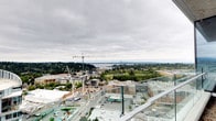 This rooftop terrace is positioned at a commanding elevation, providing an expansive, unobstructed panorama that stretches across a dynamic urban landscape. Glass panel railings line the perimeter, ensuring safety while preserving uninterrupted sightlines in every direction. The open-air design allows occupants to take in the full breadth of the surroundings, from active street-level development and mid-rise buildings in the foreground to a dense tree canopy and a shimmering body of water in the distance. On clear days, a downtown skyline is visible on the far horizon, adding a striking focal point to the view. The terrace's elevated position places it well above neighboring structures, reinforcing a sense of openness beneath the wide Pacific Northwest sky. The overall ambiance is one of urban energy tempered by natural scenery, making the space well-suited for both quiet observation and social gatherings. Its straightforward, modern design keeps the focus firmly on the remarkable surrounding environment.