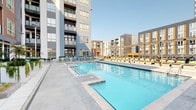 Located at 650 E. Stonewall St. in Charlotte, NC, this amenity space offers residents a well-appointed fitness center and a resort-style outdoor pool area set within a contemporary urban community. The fitness center is thoughtfully designed with a bold teal accent ceiling, white walls, and a distinctive hexagonal-patterned carpet in shades of yellow and gray that adds a modern, energetic aesthetic to the space. Large floor-to-ceiling windows flood the room with natural light while offering views of the surrounding Charlotte skyline and neighborhood. The facility is fully equipped with a comprehensive range of strength and cardio equipment, including cable machines, weight benches, a multi-station strength system, treadmills, and a full rack of free weights. Functional training amenities such as TRX suspension straps and medicine balls round out the offerings, while wall-mounted mirrors and warm wood-paneled accent walls lend a polished, upscale feel. Ceiling fans ensure comfortable airflow throughout. The adjacent outdoor pool deck features a generously sized swimming pool with a connected spa, surrounded by a clean concrete surround lined with rows of lounge chairs in yellow and gray. The pool area is framed by the community's multi-story modern residential towers, creating a vibrant and social outdoor retreat in the heart of Charlotte.