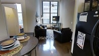 This single-floor urban space showcases a thoughtfully arranged open-concept layout that seamlessly blends the living and dining areas. The polished concrete floors serve as a defining architectural feature, reflecting light and lending an industrial-modern character to the interior. A large, multi-pane window anchors the living area, drawing in generous natural light and offering views of the surrounding cityscape, enhancing the sense of openness despite the compact footprint.

The living area is centered around dark leather seating arranged on a bold black-and-white geometric area rug, creating a striking visual contrast against the light-toned walls. A ceiling fan above provides both function and style. An entertainment unit with a flat-screen television is positioned directly beneath the window, while wall-mounted shelving adds decorative and practical storage.

Adjacent to the living space, a round dining table with a dark granite-style surface provides an intimate setting for meals. The consistent use of dark tones in the furniture and finishes gives the space a cohesive, contemporary aesthetic with an urban edge.