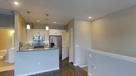 This single-floor space showcases a thoughtfully designed open-concept layout, anchored by a contemporary kitchen that serves as the focal point. The kitchen is outfitted with stainless steel appliances, including a French door refrigerator with an in-door water and ice dispenser, a built-in microwave, and a gas range with a tile backsplash. White shaker-style cabinetry lines the walls, providing ample storage, while a central island with a light grey countertop offers additional workspace and casual dining potential. Pendant lights hang above the island, adding focused illumination and a modern aesthetic touch, complemented by recessed ceiling lights throughout. The floors are finished in rich, dark hardwood, creating a warm contrast against the neutral grey-toned walls and white trim. A staircase railing is visible toward the right side of the space, suggesting access to another level or entry point. The clean lines, neutral color palette, and modern fixtures contribute to an overall ambiance that feels fresh, functional, and move-in ready.