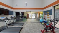 Located at 2900 Perimeter Pkwy in Augusta, GA, this fitness center is a comprehensively outfitted facility designed to support a wide range of workout routines. The space is defined by its bold, distinctive wall design — wide horizontal bands of orange and steel blue against a neutral background — that lend the interior a modern, energetic character. Soaring ceilings amplify the sense of openness throughout the main workout area.

The cardio zone is well-stocked with a row of TRUE-brand treadmills, upright and recumbent bikes, and elliptical trainers, all positioned to take advantage of large windows that frame views of the surrounding landscaping. Wall-mounted televisions are conveniently placed throughout this section for entertainment during workouts.

The strength and free weight area features a tiered dumbbell rack, adjustable weight benches, and a cable/functional training machine with TRX suspension straps, providing versatile options for resistance training. Exercise balls and specialty equipment are also available for functional and recovery work.

A notable highlight of the facility is the enclosed indoor golf simulator bay, complete with a green turf mat and a full projection screen, offering a unique recreational amenity within the fitness space. The flooring throughout consists of durable, patterned carpet tile suited for high-traffic athletic use. Glass partitions and an open layout connect the various zones cohesively.