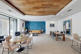 Located at 2800 Kirby Dr in Houston's vibrant Upper Kirby neighborhood, this single-floor space serves as a well-appointed communal lounge and workspace. The interior is defined by a striking teal and gold geometric accent wall that anchors the main lounge area, where a tan leather sofa and a black armchair create a welcoming conversation nook. Large floor-to-ceiling sliding glass doors flood the space with natural light and open directly onto an outdoor patio, seamlessly blending indoor and outdoor living. A dedicated workstation area features built-in computer desks set against the signature teal accent wall, offering a functional workspace within the stylish setting. The space transitions between carpet and warm hardwood flooring, delineating the various zones. A raised dining area with a round wooden table and upholstered chairs sits beneath a refined tray ceiling detailed with natural wood paneling, adding warmth and architectural interest overhead. A private conference nook provides a more intimate setting with a solid wood table and executive seating. Curated contemporary artwork is displayed throughout, enhancing the modern, design-forward ambiance of this versatile and inviting space.