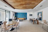 Located at 2800 Kirby Dr in Houston's vibrant Upper Kirby neighborhood, this single-floor space serves as a well-appointed communal lounge and workspace. The interior is defined by a striking teal and gold geometric accent wall that anchors the main lounge area, where a tan leather sofa and a black armchair create a welcoming conversation nook. Large floor-to-ceiling sliding glass doors flood the space with natural light and open directly onto an outdoor patio, seamlessly blending indoor and outdoor living. A dedicated workstation area features built-in computer desks set against the signature teal accent wall, offering a functional workspace within the stylish setting. The space transitions between carpet and warm hardwood flooring, delineating the various zones. A raised dining area with a round wooden table and upholstered chairs sits beneath a refined tray ceiling detailed with natural wood paneling, adding warmth and architectural interest overhead. A private conference nook provides a more intimate setting with a solid wood table and executive seating. Curated contemporary artwork is displayed throughout, enhancing the modern, design-forward ambiance of this versatile and inviting space.