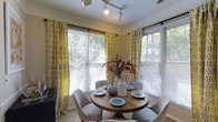 This 2-bedroom, 2-bathroom residence offers a thoughtfully designed single-floor layout that balances comfort and style. The dining area is a standout feature, bathed in abundant natural light streaming through multiple large windows dressed with cheerful yellow patterned curtains and white horizontal blinds. A rustic round wooden dining table surrounded by upholstered chairs anchors the space, creating an inviting setting for meals and gatherings. Track lighting overhead provides flexible illumination, complementing the warm, neutral wall tones throughout. Lush greenery visible through the windows adds a sense of connection to the outdoors, enhancing the bright and airy ambiance of the interior. The overall aesthetic is warm and welcoming, with a blend of natural textures and soft furnishings that contribute to a cohesive, livable atmosphere. With two well-appointed bedrooms and two full bathrooms, the layout is well-suited for a variety of lifestyles, offering both private retreats and shared living areas that feel open and connected. The attention to detail in the finishes and décor reflects a space designed for both everyday functionality and comfortable living.
