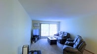 This single-floor space centers around a comfortable and functional living room. The room features wall-to-wall carpeting that adds warmth underfoot, paired with smooth, light-colored walls that create a clean, neutral backdrop. A large sliding glass door dominates one wall, fitted with vertical blinds that allow for adjustable natural light and provide a view of the surrounding outdoor greenery. The living area is furnished with a generously sized reclining sofa and matching recliner chairs, arranged to face a low-profile media console housing a flat-screen television. A rectangular coffee table sits at the center of the seating arrangement, offering a practical surface for everyday use. A baseboard heating unit runs along the exterior wall beneath the sliding door, indicating a practical and efficient heating setup throughout the space. Framed wall art adds a personal touch above the media console. The overall ambiance is casual and inviting, with an emphasis on comfortable, everyday living. The open layout of the living area allows for easy movement and versatile furniture arrangement, making it a practical and livable space suited for relaxing and entertaining.