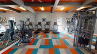 This fitness center makes an immediate visual impression with its bold, energetic design. The flooring features a striking checkered pattern in orange, teal, and gray, while the ceiling carries the same vibrant palette — with orange-painted recessed panels framed by exposed gray structural beams. Ceiling fans and flush-mounted fluorescent lighting ensure the space remains bright and comfortable throughout.

The facility is generously laid out across a single open floor, providing ample room for a full range of exercise equipment. The cardio zone includes multiple treadmills equipped with individual screens, an elliptical trainer, a stationary recumbent bike, and a stair climber. A dedicated strength training area features a multi-station cable machine, a power rack, and a full dumbbell rack ranging across various weights. A flat-screen TV and a digital clock are mounted on the wall for convenience during workouts.

Large French doors and multiple windows flood the space with natural light and offer views to the outdoors, creating an open, airy atmosphere. Full-length mirrors line one wall, visually expanding the room and supporting proper form during training. Framed artwork is displayed throughout, adding a polished, finished feel to the space. The overall ambiance is energetic and motivating, combining functional design with a distinctly modern aesthetic.