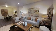 This single-floor space showcases a thoughtfully designed living area that blends comfort and style through a cohesive neutral palette of warm grays, taupes, and earthy tones. The living room serves as the focal point, centered around a light gray upholstered sofa accented with burnt orange and patterned throw pillows, complemented by a woven rattan armchair that adds natural texture. A large, framed abstract painting in muted golds, blushes, and grays anchors the main wall, lending an artistic and sophisticated touch to the space.

A rustic wooden coffee table with metal legs sits atop a cowhide rug layered over a natural fiber area rug, adding depth and warmth to the flooring. Decorative ceramic vases and curated accessories on the coffee table enhance the styled aesthetic. Ambient lighting is provided by table lamps with warm-toned shades, creating an inviting atmosphere throughout.

An open passageway connects the living area to a bedroom, which is visible in the background and features similarly warm, layered bedding and a classic table lamp. The overall ambiance of the space is cozy, elegant, and well-coordinated, reflecting a cohesive interior design sensibility.
