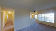 This single-floor residence offers a clean, open layout designed to maximize both comfort and functionality. The living area is finished with plush wall-to-wall carpeting and is bathed in natural light streaming through large windows, creating a warm and inviting atmosphere. Crown molding runs along the ceiling, adding a refined architectural detail that elevates the overall aesthetic of the space.

A defining feature of the layout is the pass-through kitchen counter, which connects the kitchen to the living area seamlessly. A pendant light hangs above the pass-through, providing focused illumination while reinforcing the connection between the two spaces. The kitchen itself benefits from the same abundance of natural light visible throughout the residence.

A hallway visible from the main living area leads to the remaining rooms, suggesting a thoughtful floor plan that offers both open communal spaces and more private areas. The walls are finished in a soft, neutral tone that complements the carpeted floors and contributes to the light, airy feel of the home. HVAC venting is integrated discreetly into the ceiling, ensuring year-round climate comfort without compromising the clean visual lines of the interior. Overall, this residence presents a well-maintained, functional living environment with classic finishing touches.