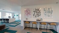 Located at 215 107th Street Circle East in Bradenton, Florida, this single-floor community amenity space is designed with a modern, vibrant aesthetic that encourages both collaboration and relaxation. The interior is immediately defined by a bold chevron-patterned carpet in teal, gray, and beige tones that flows throughout the entire space, tying each zone together cohesively.

The open layout encompasses several distinct functional areas. A lounge zone features a large teal sectional sofa accented with yellow throw pillows, complemented by rounded ottoman seating in coordinating teal and brown tones — all oriented toward natural light streaming in through large windows. Adjacent to this is a flexible seating cluster with modular navy, charcoal, and yellow upholstered pieces, ideal for casual gatherings.

A built-in counter-height bar along one wall provides a dedicated workspace or social perch, outfitted with warm wood and metal bar stools beneath a curated gallery of bold abstract artwork. A co-working area features desktop computer stations at a sleek white workspace, offering a productive environment with ample natural light.

Glass-walled rooms along one side of the space house a fitness facility, visible through floor-to-ceiling framed glass doors. A wall-mounted flat-screen television and thoughtfully placed accent lighting round out this well-appointed, multi-use community space.
