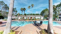 This outdoor pool area offers a resort-style retreat designed for relaxation and leisure. The centerpiece is a generously sized swimming pool with clear, blue water, framed by a well-maintained paver brick deck that provides ample space for lounging and socializing. Wrought-iron patio tables and chairs are thoughtfully arranged around the deck, offering comfortable spots to unwind poolside. Lush tropical palm trees and towering pine trees surround the area, creating a natural, shaded backdrop that enhances the atmosphere of seclusion and tranquility. Additional lounge seating is visible along the pool's perimeter, accommodating a variety of recreational needs. A low-rise building serves as an attractive backdrop, featuring a covered entryway flanked by structured landscaping and manicured greenery. The surrounding foliage and open sky contribute to a bright, airy environment that feels both expansive and welcoming. The combination of tropical landscaping, quality hardscaping, and well-appointed furnishings makes this pool area a standout amenity, ideal for enjoying warm, sunny days in a serene outdoor setting.